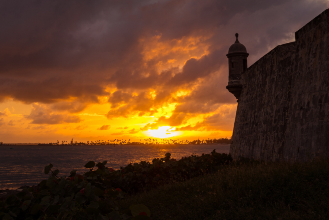 Sunset over a coastal landscape with a silhouette of a stone wall and tower.