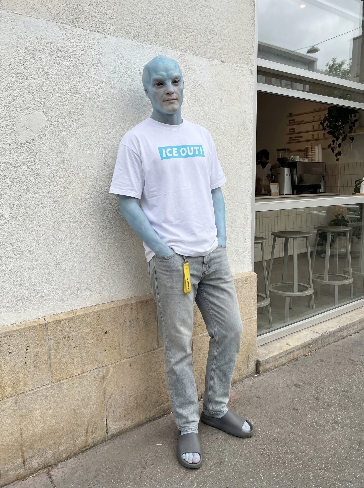Person with blue skin wearing a white t-shirt with 'ICE OUT' logo and gray pants, standing against a building.