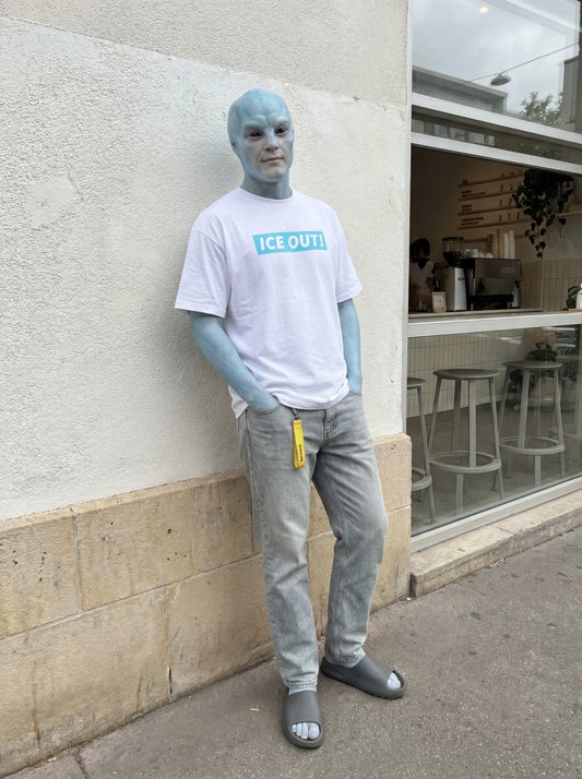 Person with blue skin wearing a white t-shirt with 'ICE OUT' logo and gray pants, standing against a building.